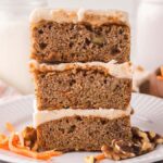 Three frosted carrot cake bars stacked on a plate, with walnuts and shredded carrots beside these delicious carrot cake bars.