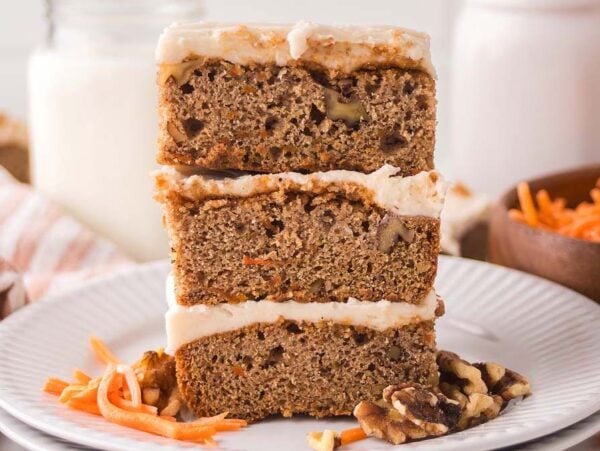 Three frosted carrot cake bars stacked on a plate, with walnuts and shredded carrots beside these delicious carrot cake bars.