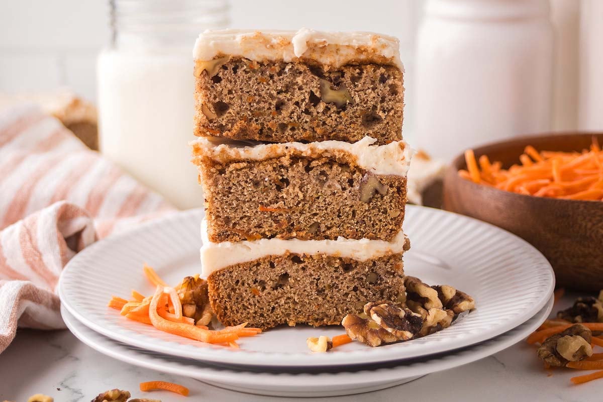 Three frosted carrot cake bars stacked on a white plate, with walnuts and shredded carrots beside these moist carrot cake bars.