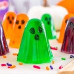 Colorful, ghost jello shots shaped like playful spirits with black eyes and mouths, surrounded by Halloween-themed sprinkles.