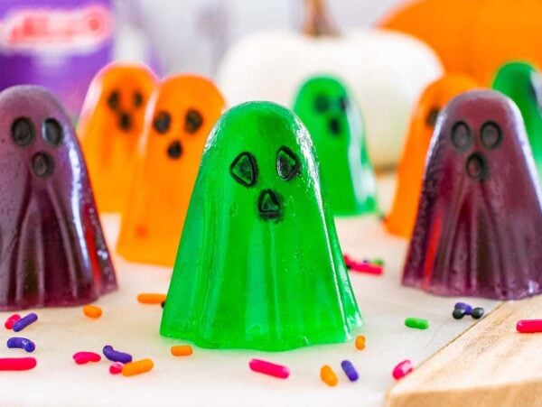 Colorful, ghost jello shots shaped like playful spirits with black eyes and mouths, surrounded by Halloween-themed sprinkles.