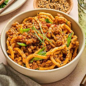 Bowl of spicy Miso-Chili Tempeh Noodles with ground meat and green onions, beside a drink, chopsticks, and spice dishes.