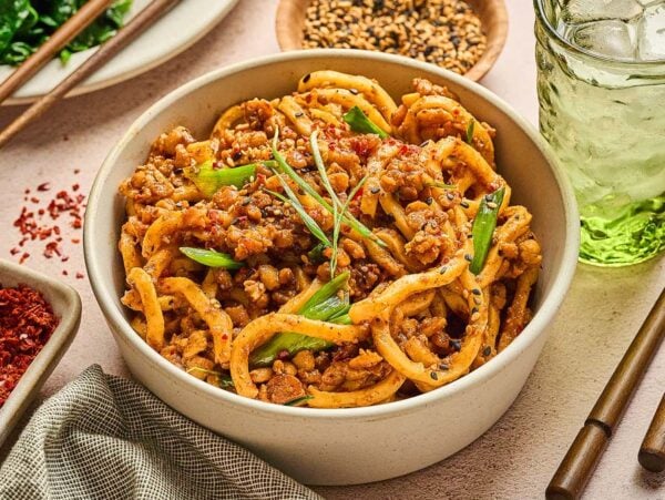 Bowl of spicy Miso-Chili Tempeh Noodles with ground meat and green onions, beside a drink, chopsticks, and spice dishes.