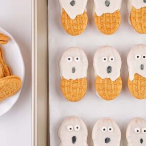 Nutter Butter Ghost Cookies decorated with white icing and candy eyes, surrounded by fake spiders for a spooky treat.