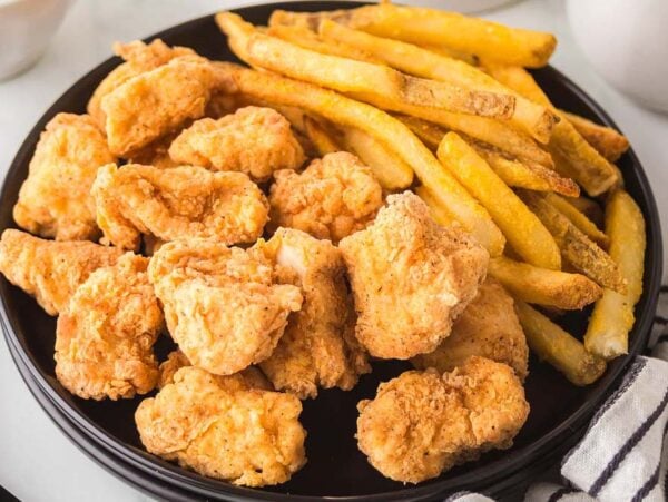 A black plate with crispy popcorn chicken and golden French fries sits beside a striped napkin.