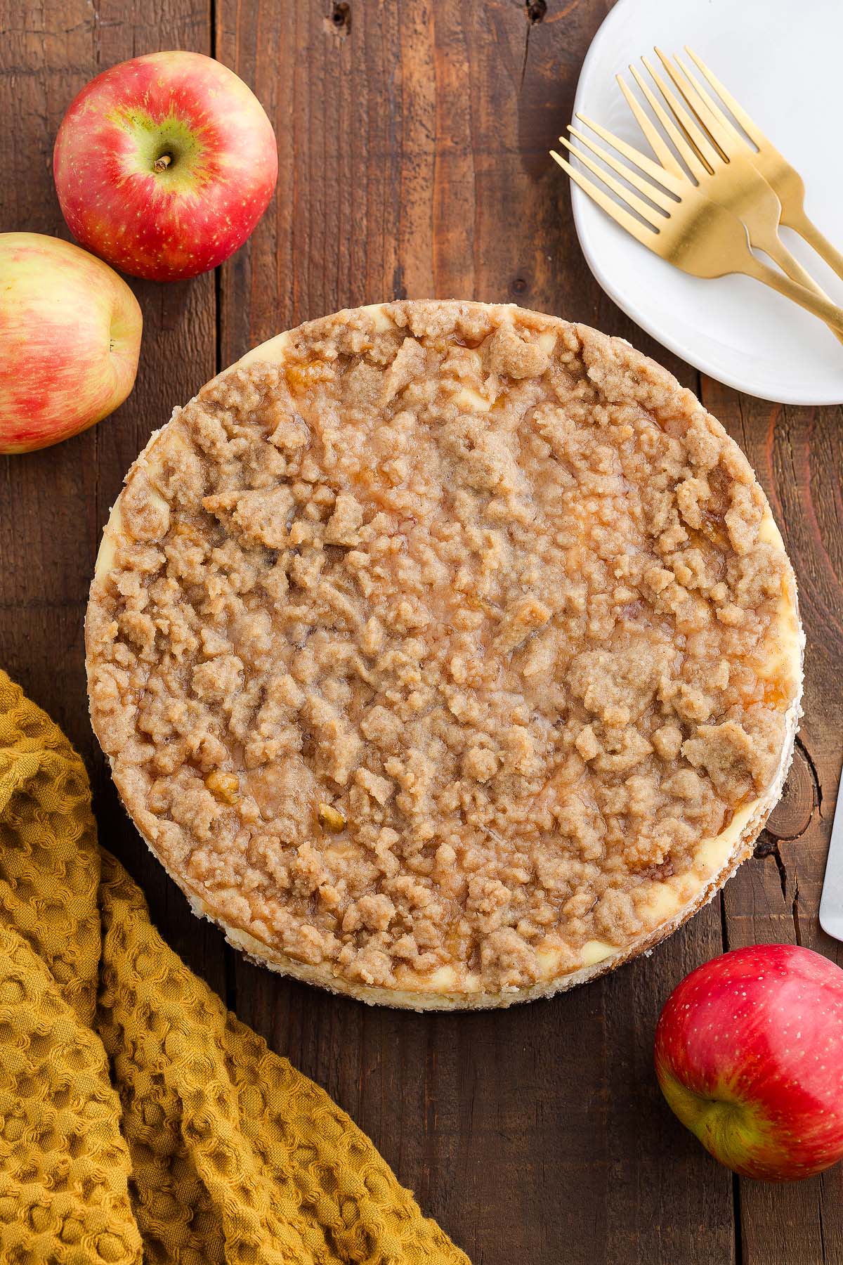 A whole apple pie cheesecake sits on a wooden table with apples, plates, forks, and a yellow cloth nearby.