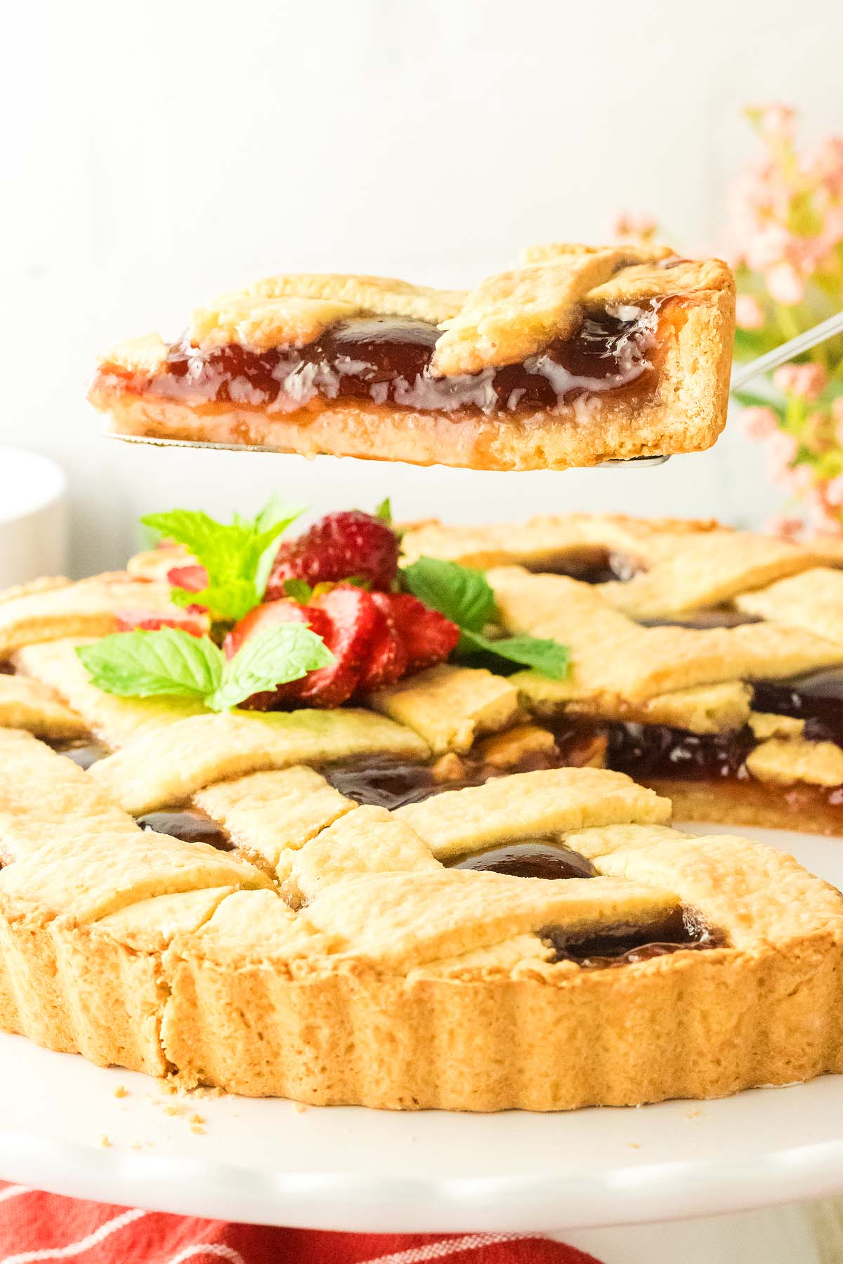 A slice of lattice-topped crostata di marmellata is being lifted from a whole tart, beautifully garnished with strawberries and mint.