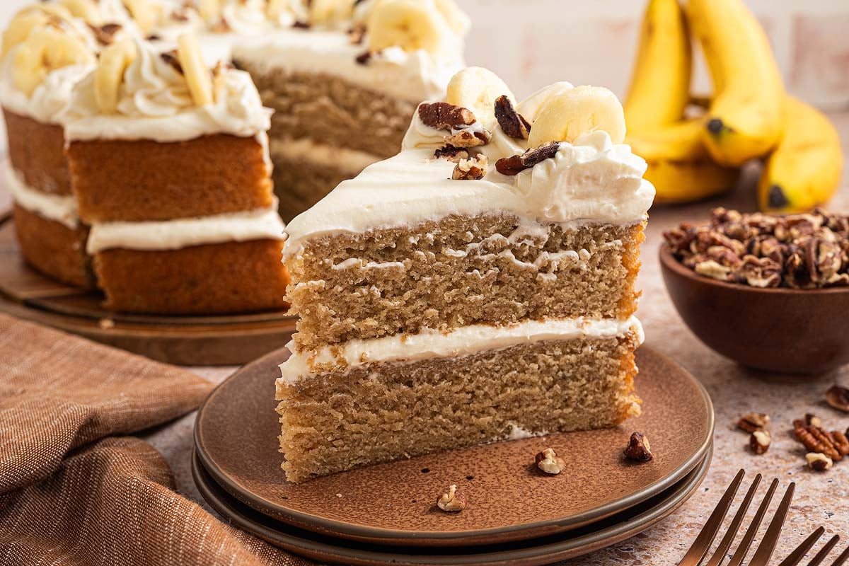 A slice of banana cake with cream frosting sits on a plate, surrounded by fresh bananas and nuts in the background.