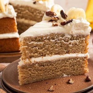 A slice of Banana Cake with cream frosting and pecans, served with fresh bananas in the background.