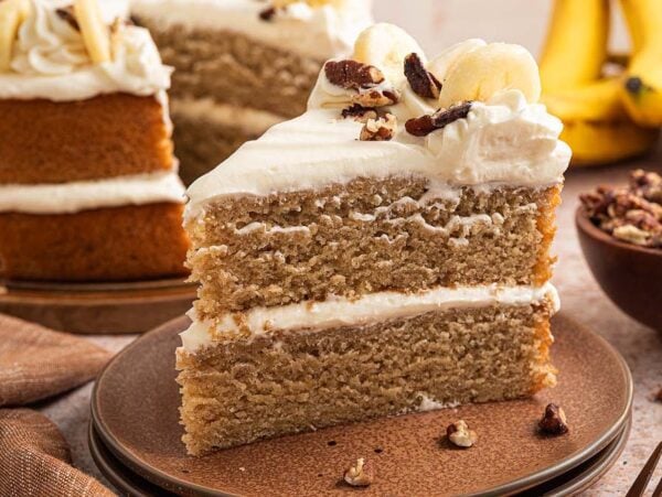 A slice of Banana Cake with cream frosting and pecans, served with fresh bananas in the background.