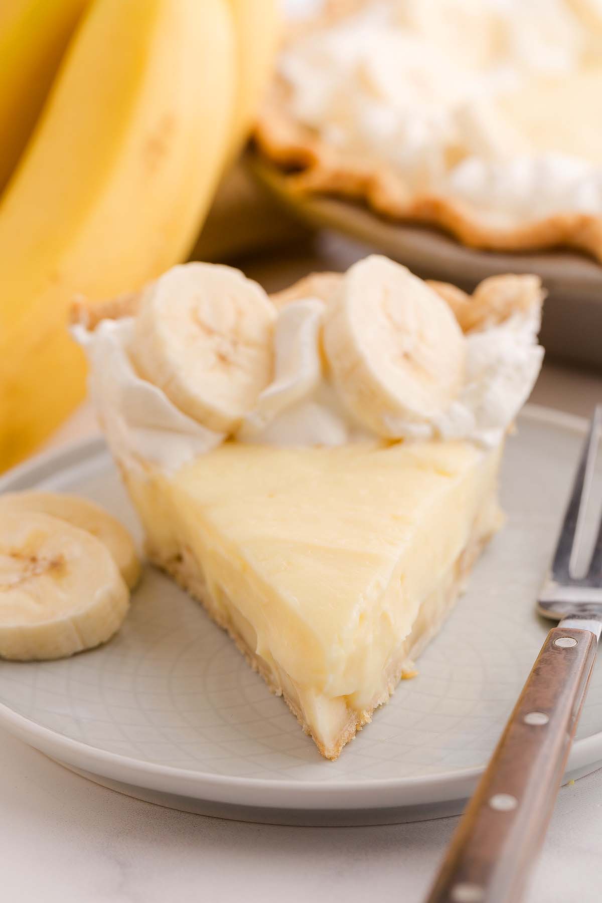 A decadent slice of banana cream pie topped with whipped cream and fresh banana slices sits on a plate, ready to enjoy with a fork beside it.