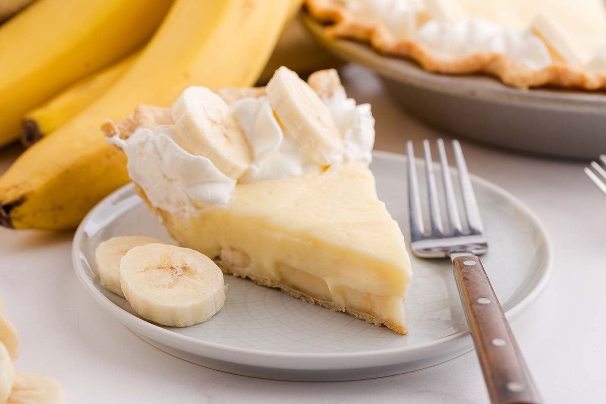 A slice of banana cream pie topped with whipped cream and fresh banana slices, served on a plate with a fork.
