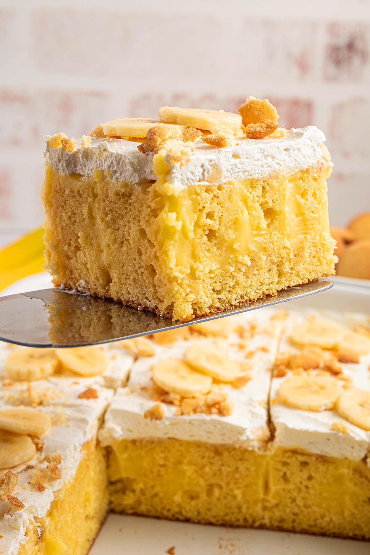 A slice of banana pudding poke cake with whipped topping and banana slices is being lifted from a tray, showcasing the creamy layers and irresistible flavor of this classic dessert.