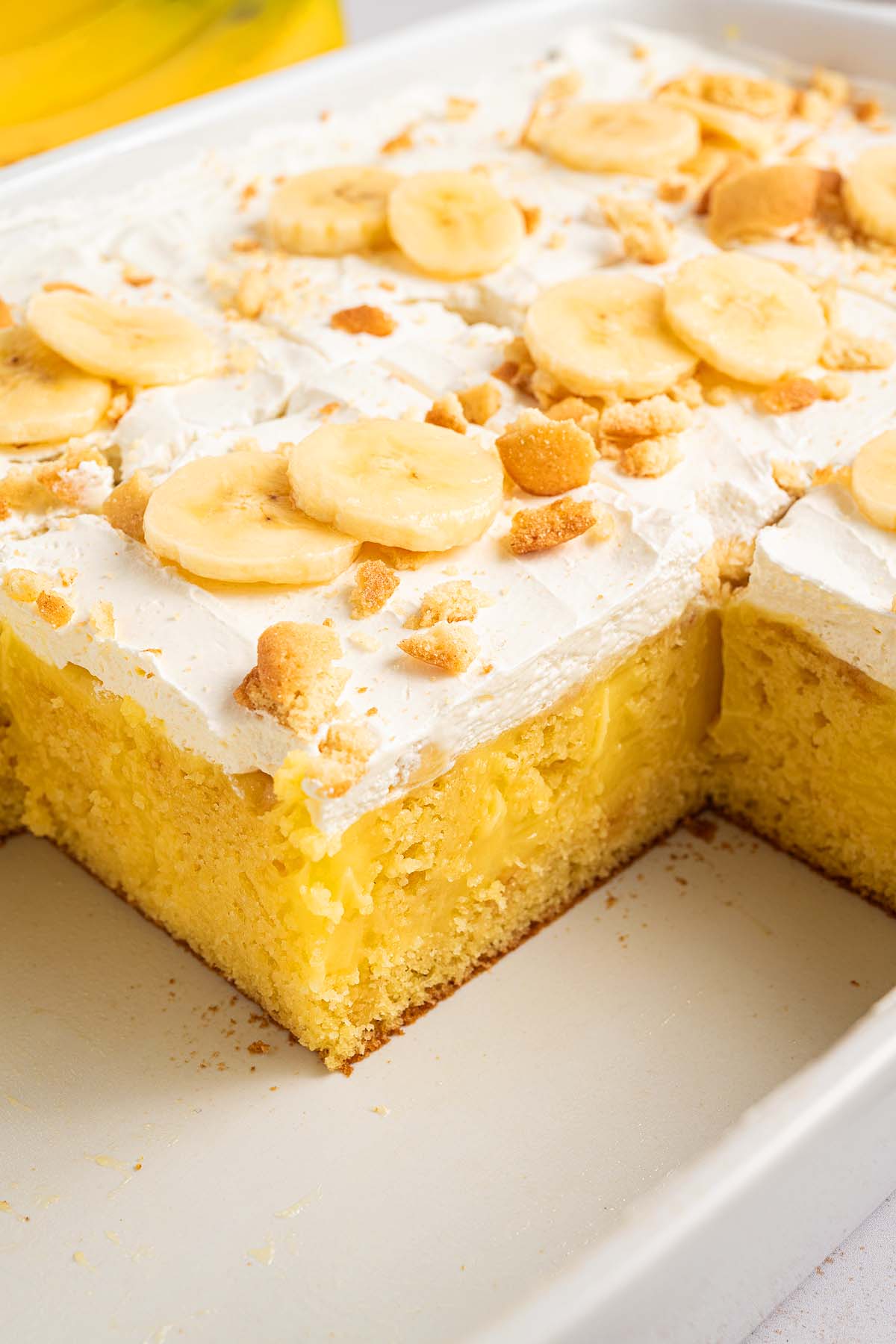 Banana pudding poke cake made with yellow cake, topped with whipped cream, banana slices, and crumbled cookies in a white baking dish.