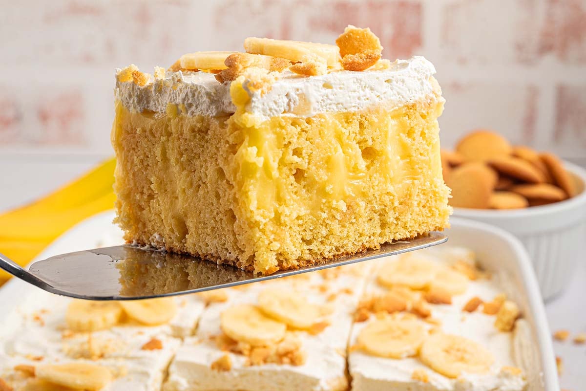 A slice of banana pudding poke cake with whipped topping and banana slices is lifted on a spatula.