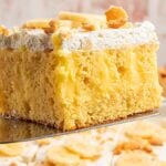 A slice of banana pudding poke cake with whipped topping and wafer pieces, held above a tray of cake.