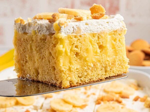 A slice of banana pudding poke cake with whipped topping and wafer pieces, held above a tray of cake.