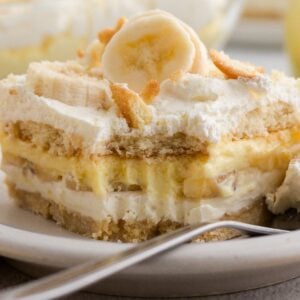 A slice of Banana Pudding dessert with whipped cream and banana slices sits on a white plate, with a fork beside it.