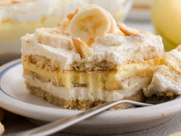A slice of Banana Pudding dessert with whipped cream and banana slices sits on a white plate, with a fork beside it.