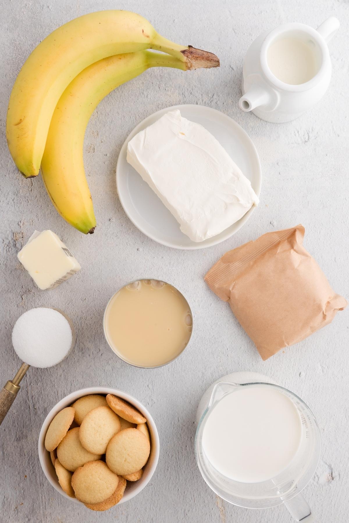 Ingredients on a light surface: bananas, cream cheese, milk, cookies, butter, sugar, condensed milk, paper packet.