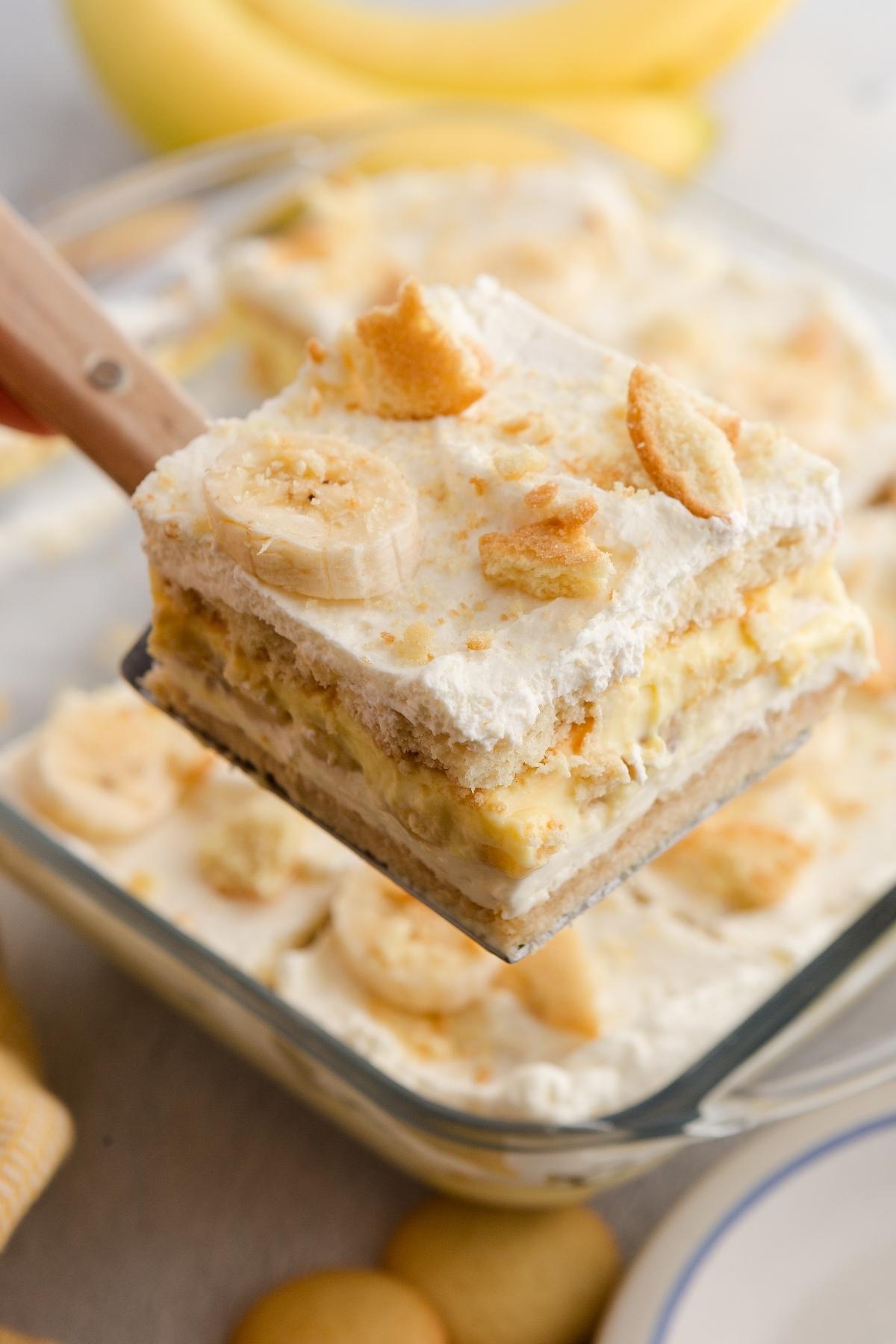 A spatula lifts a slice of banana pudding with whipped topping and cookie crumbles from a glass dish.