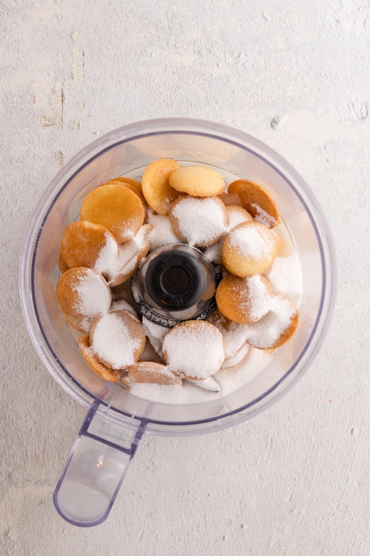 Vanilla wafers and sugar in a food processor bowl on a light-colored surface, ready to be blended.