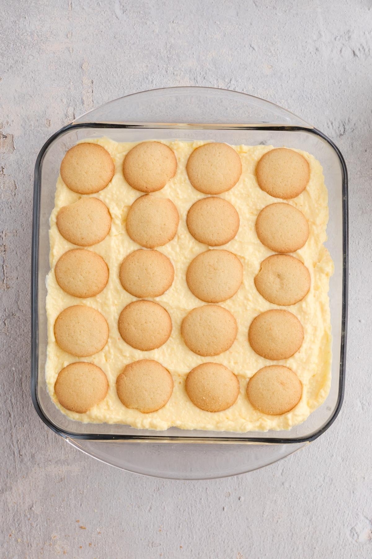 Square glass dish with creamy pudding topped with rows of round vanilla wafer cookies on a light surface.