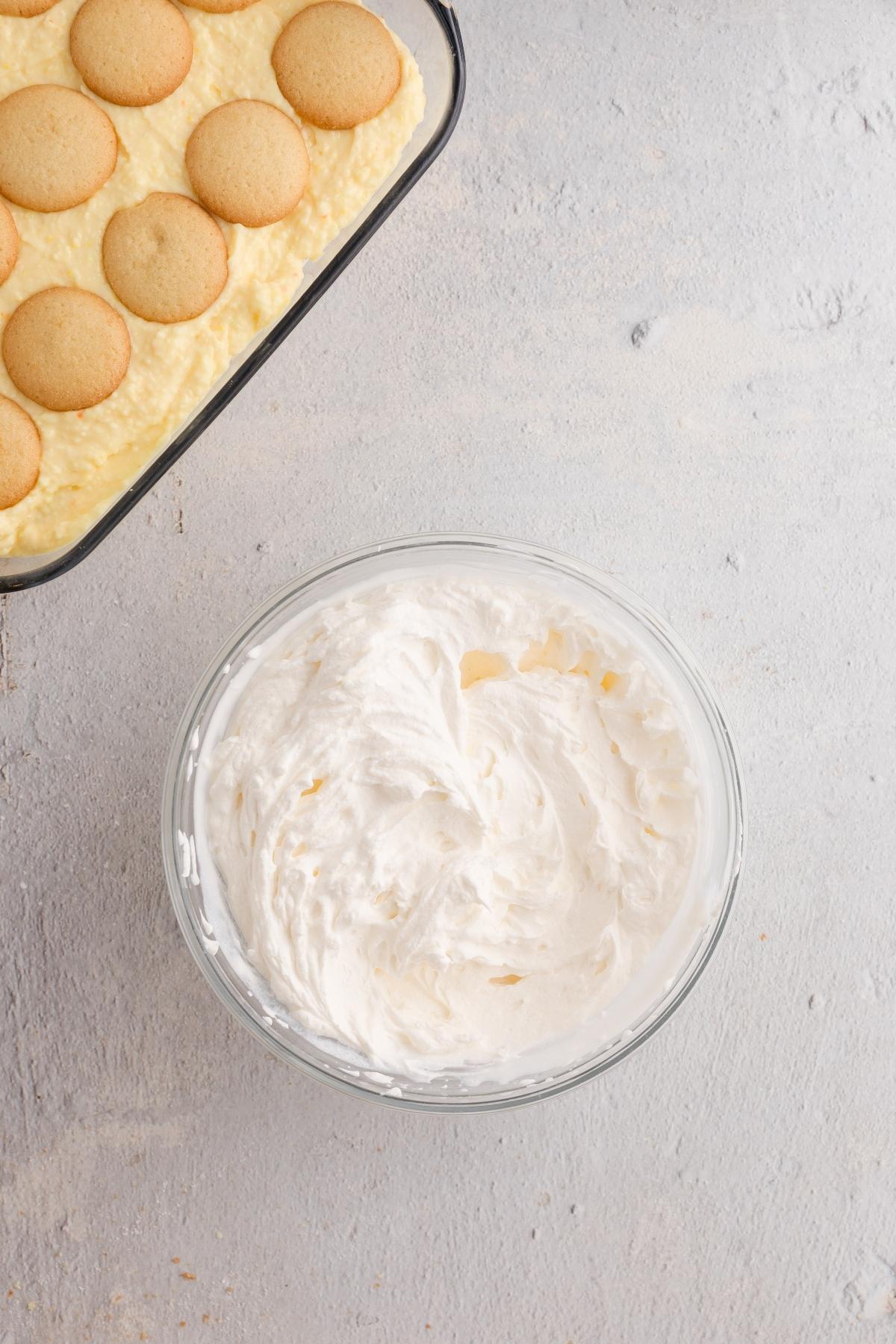 A bowl of whipped cream next to a baking dish with vanilla wafers on pudding.