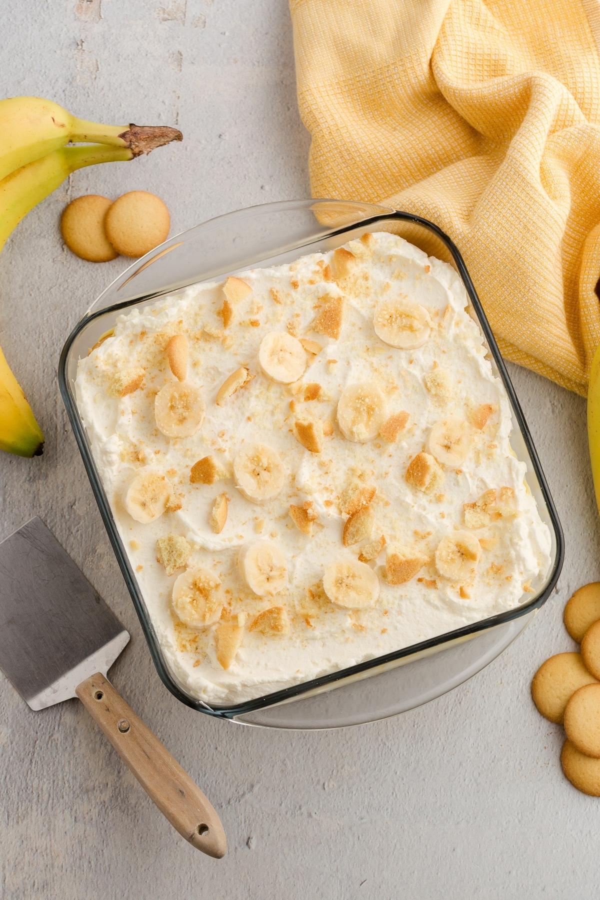 A glass dish of banana pudding topped with banana slices and vanilla wafers, next to whole bananas and cookies.
