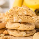 A stack of Banana Pudding Cookies with white chocolate chips, surrounded by cookie crumbs and bananas in the background.