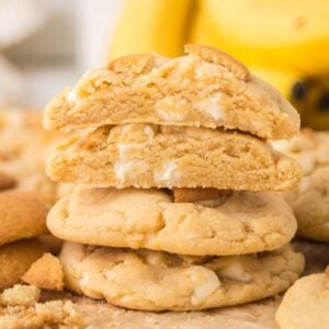 A stack of Banana Pudding Cookies with white chocolate chips, surrounded by cookie crumbs and bananas in the background.