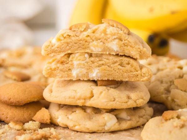 A stack of Banana Pudding Cookies with white chocolate chips, surrounded by cookie crumbs and bananas in the background.