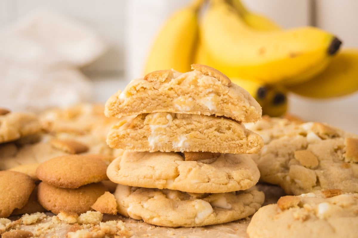 A stack of Banana Pudding Cookies with white chocolate chips, surrounded by more cookies and bananas in the background.