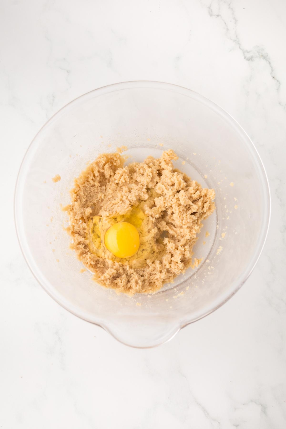 A clear bowl with creamed sugar and butter and a cracked egg on top, on a white marble surface.