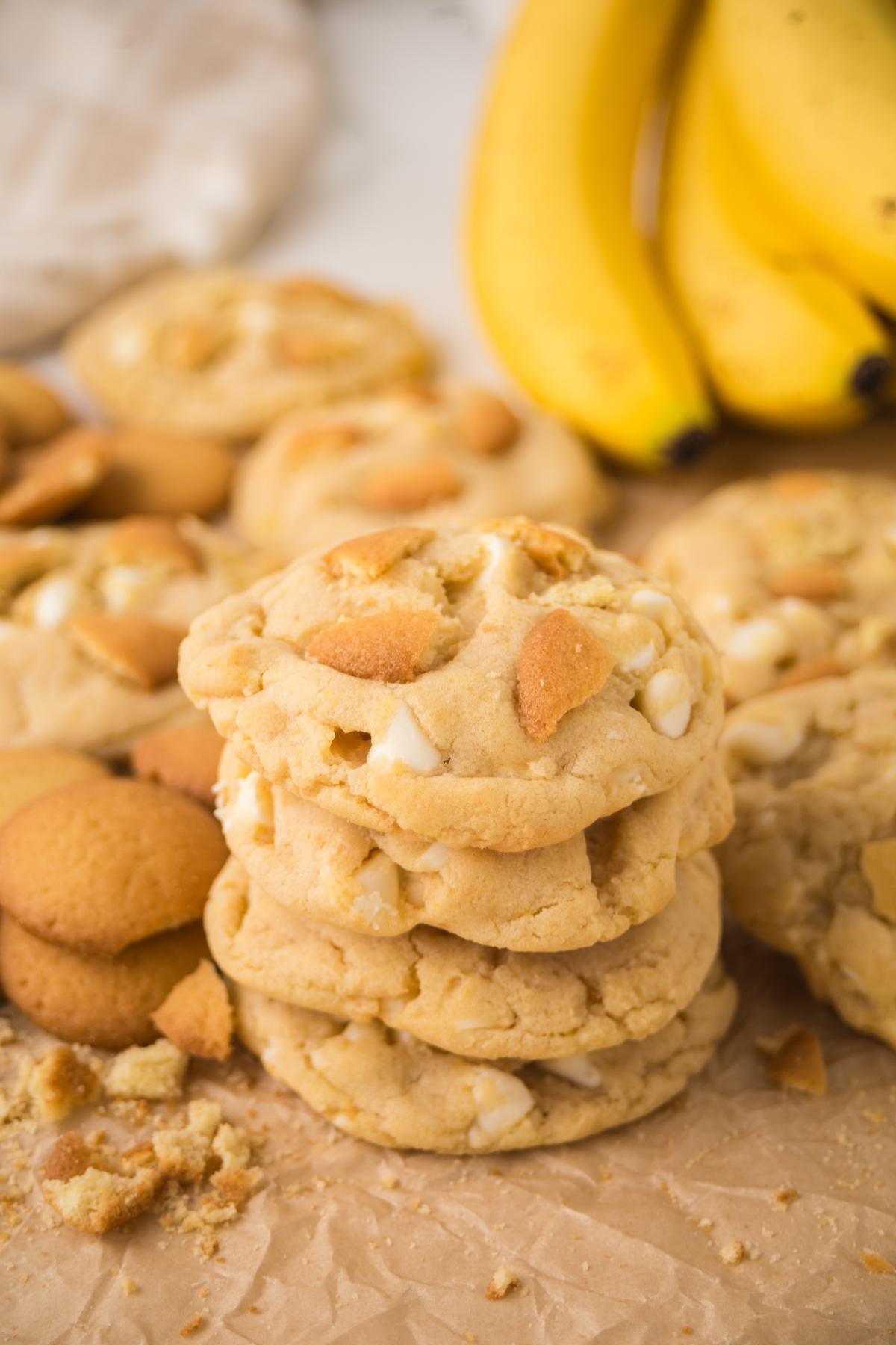 Stack of cookies with white chocolate chips and cookie pieces, bananas and more cookies in the background.