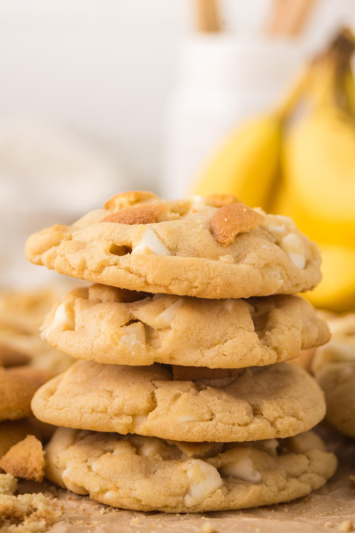 Four stacked white chocolate chip cookies with banana slices in the background.