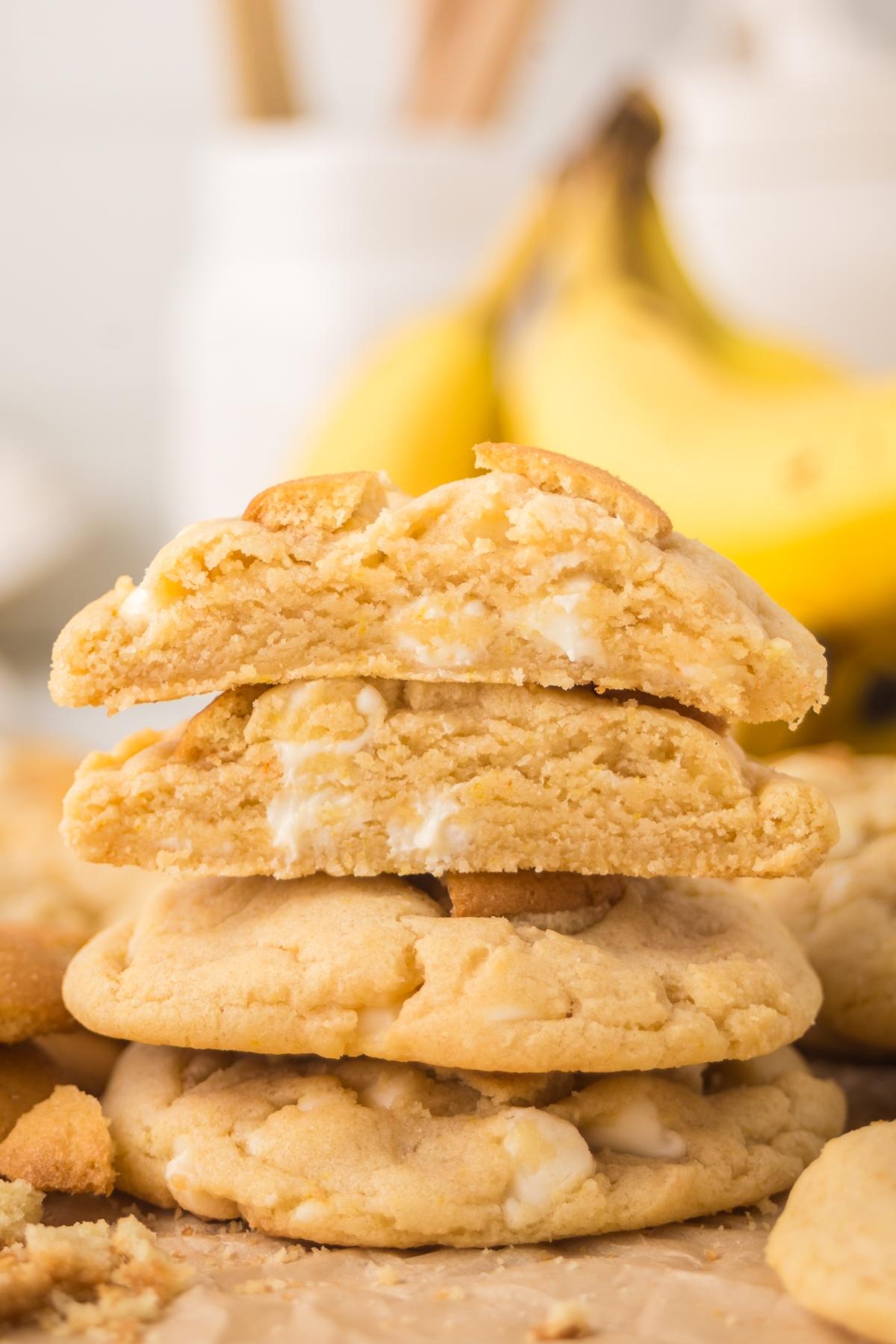 A stack of banana pudding cookies with white chocolate chips, one broken in half, with bananas in the background.