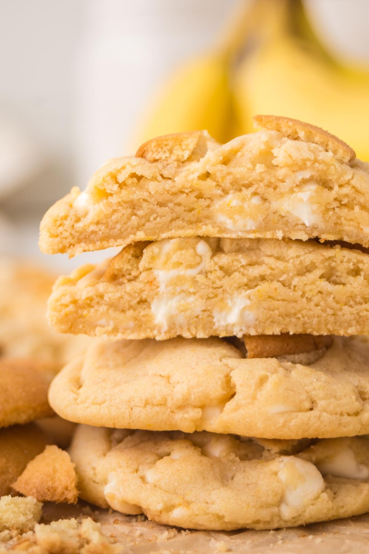 Stack of soft, golden cookies with white chocolate chips, one broken in half, with bananas in the background.