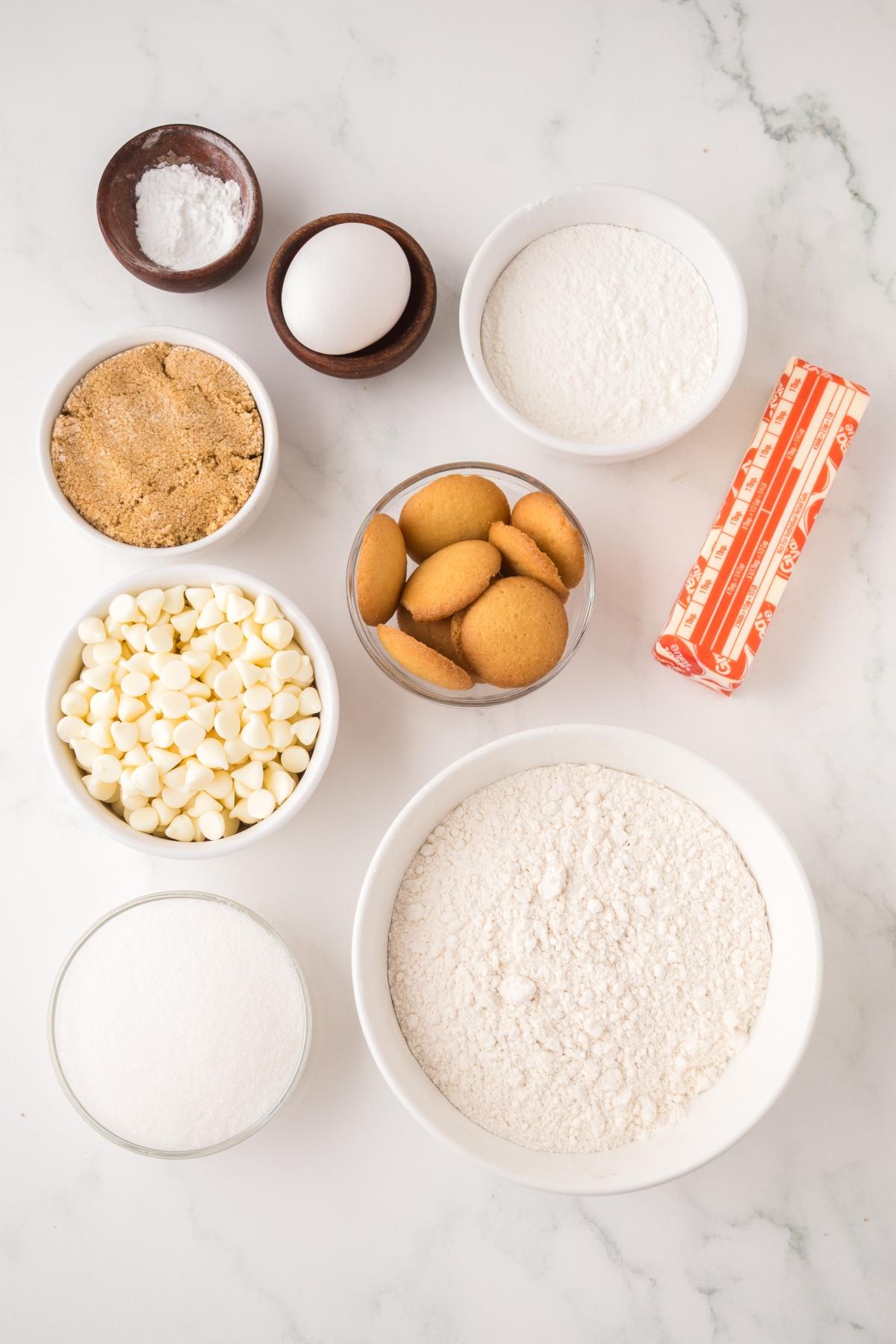 Bowls of flour, sugar, brown sugar, white chocolate chips, vanilla wafers, butter, baking powder, and an egg.