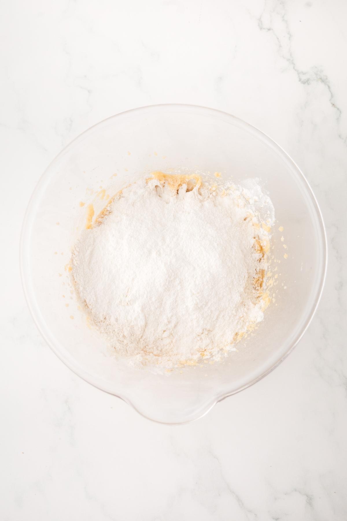 A clear mixing bowl with flour and other baking ingredients on a white marble surface.