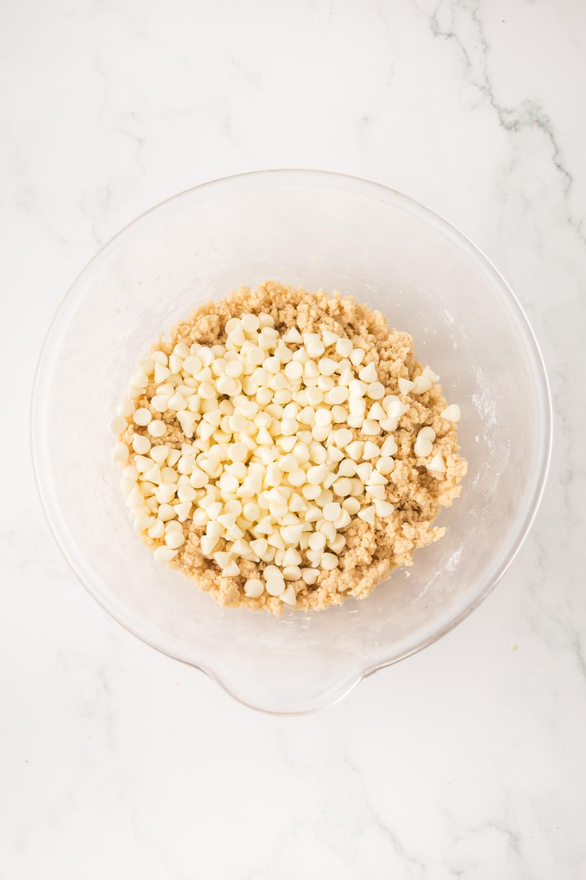 A glass bowl with cookie dough and white chocolate chips on a white marble surface.