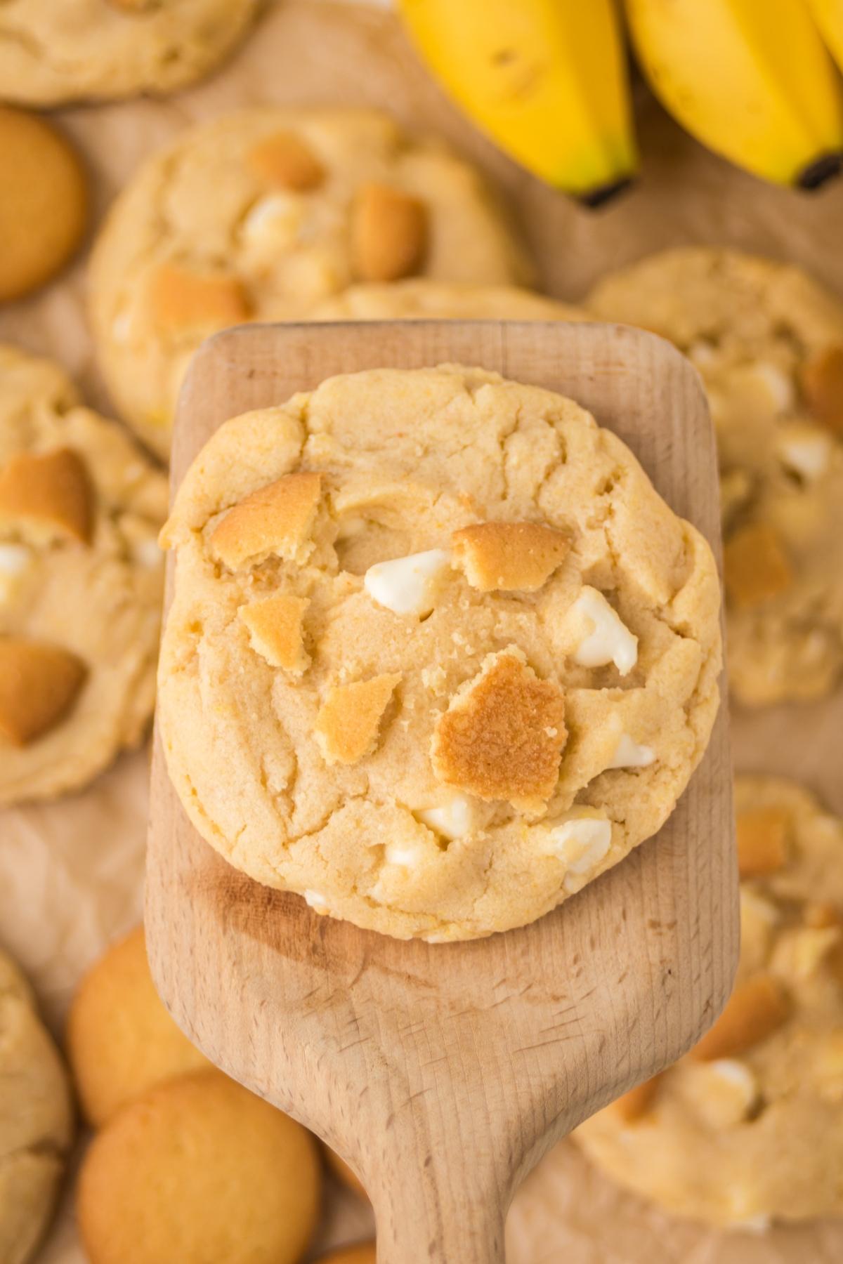 A cookie with white chocolate chips and vanilla wafer pieces on a wooden spatula, bananas in the background.