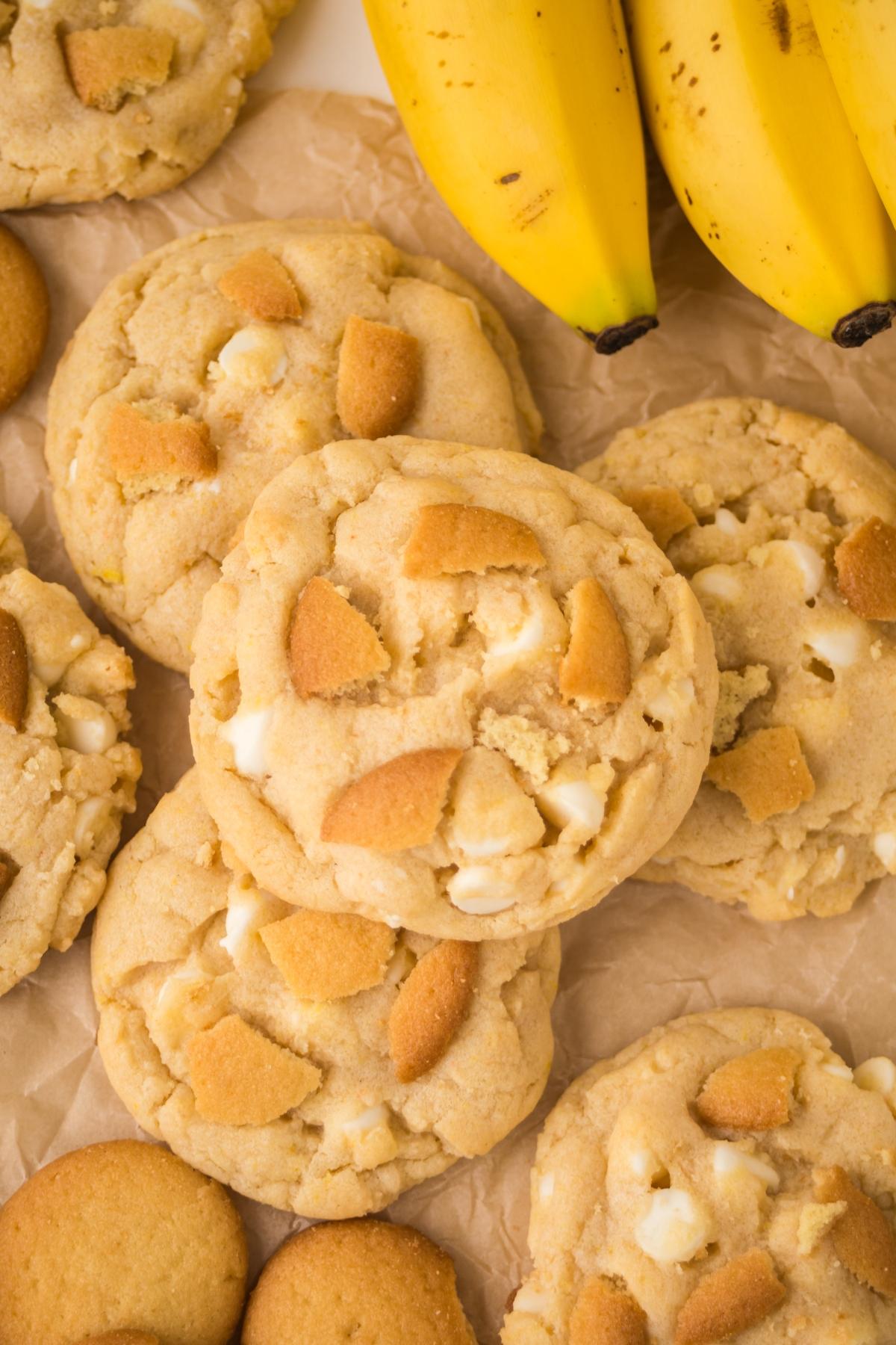 Cookies with vanilla wafer pieces on parchment paper next to a bunch of ripe bananas.