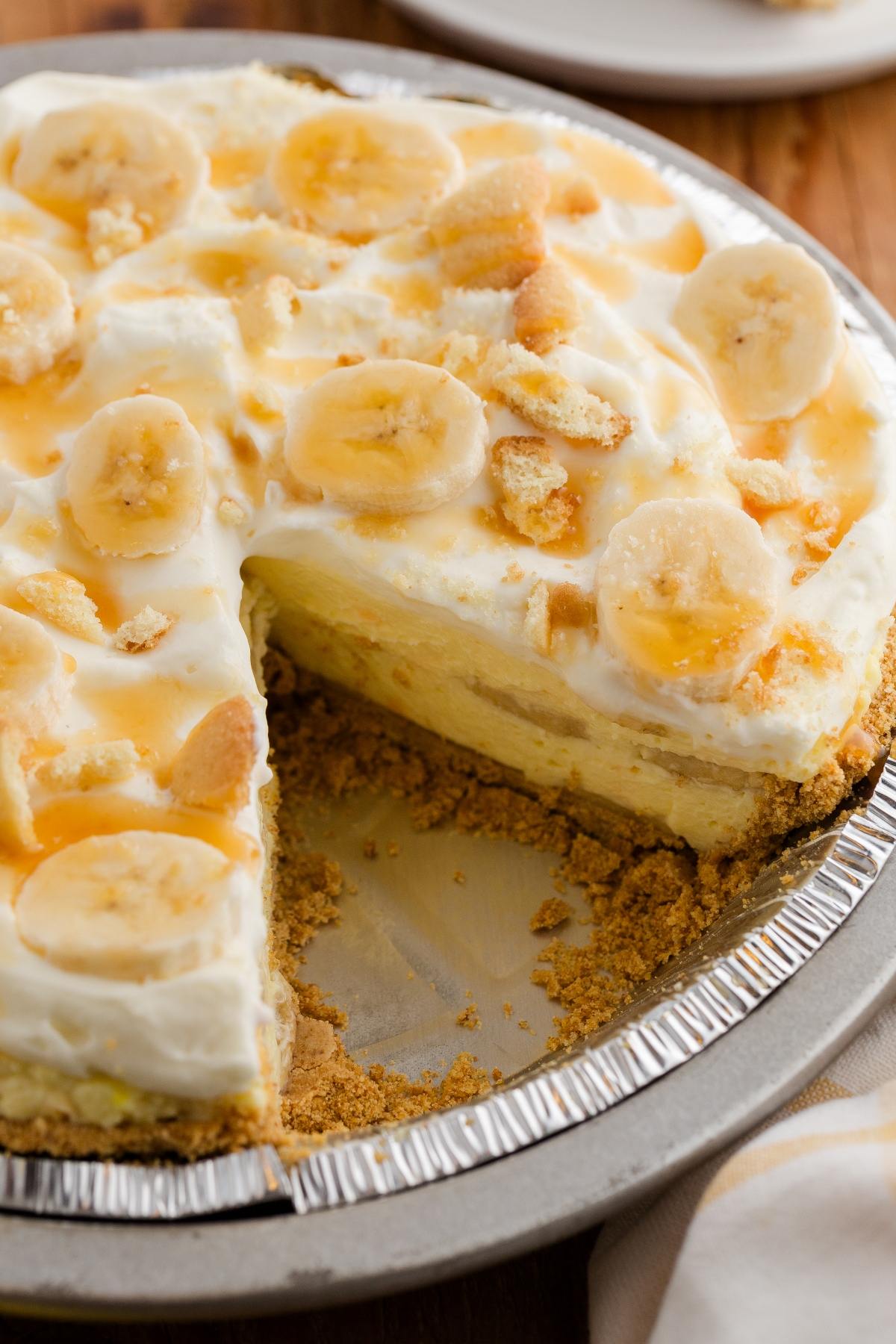 A banana pudding pie with whipped topping, banana slices, and a slice missing, in a metal pie tin.