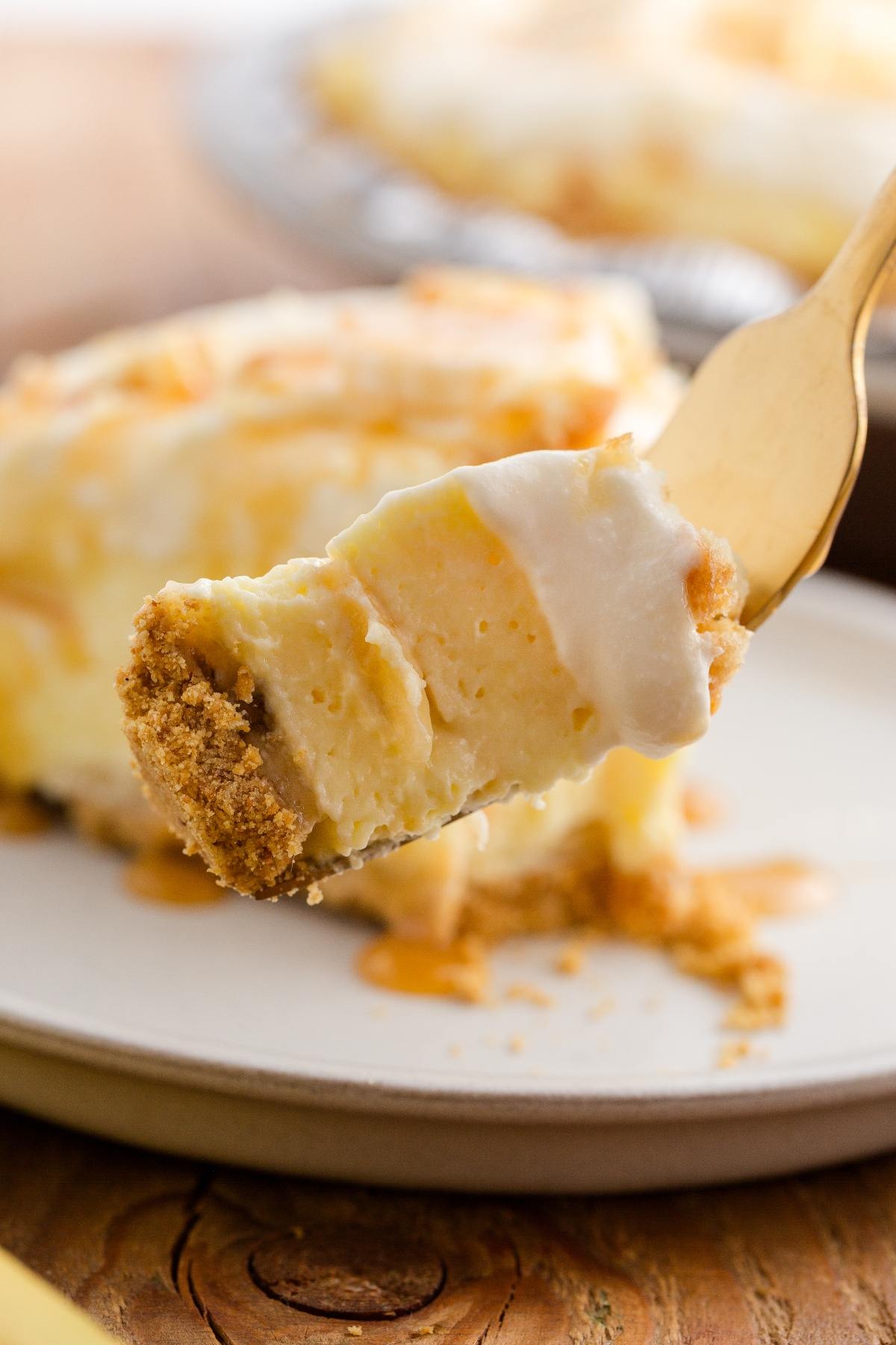 A fork holds a creamy slice of pie with a graham cracker crust above a white plate.