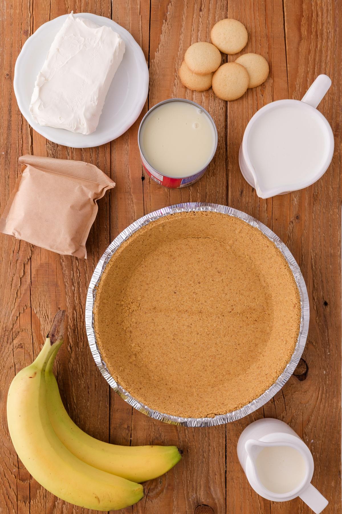 Pie crust, bananas, cream cheese, cookies, sweetened milk, milk, and a brown pouch on a wooden surface.