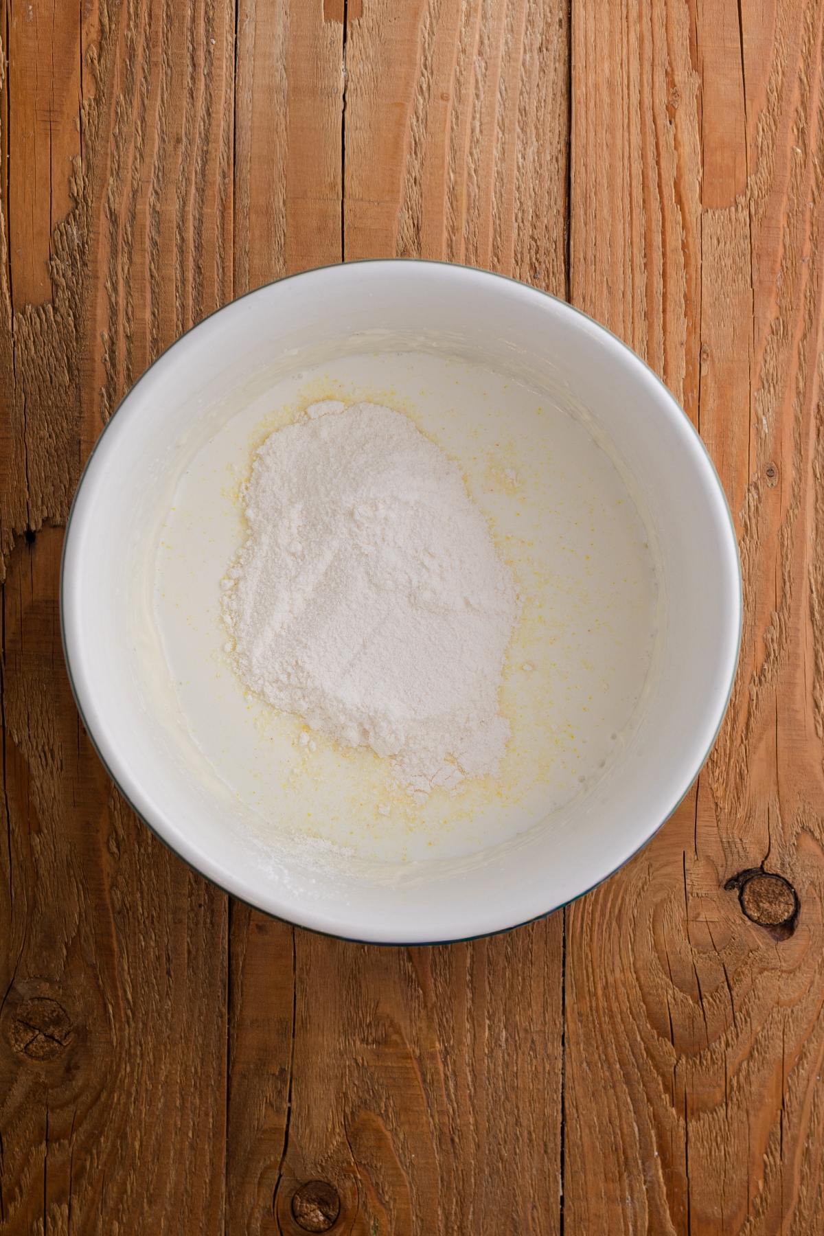 White bowl with flour and liquid mixture on a wooden surface, viewed from above.