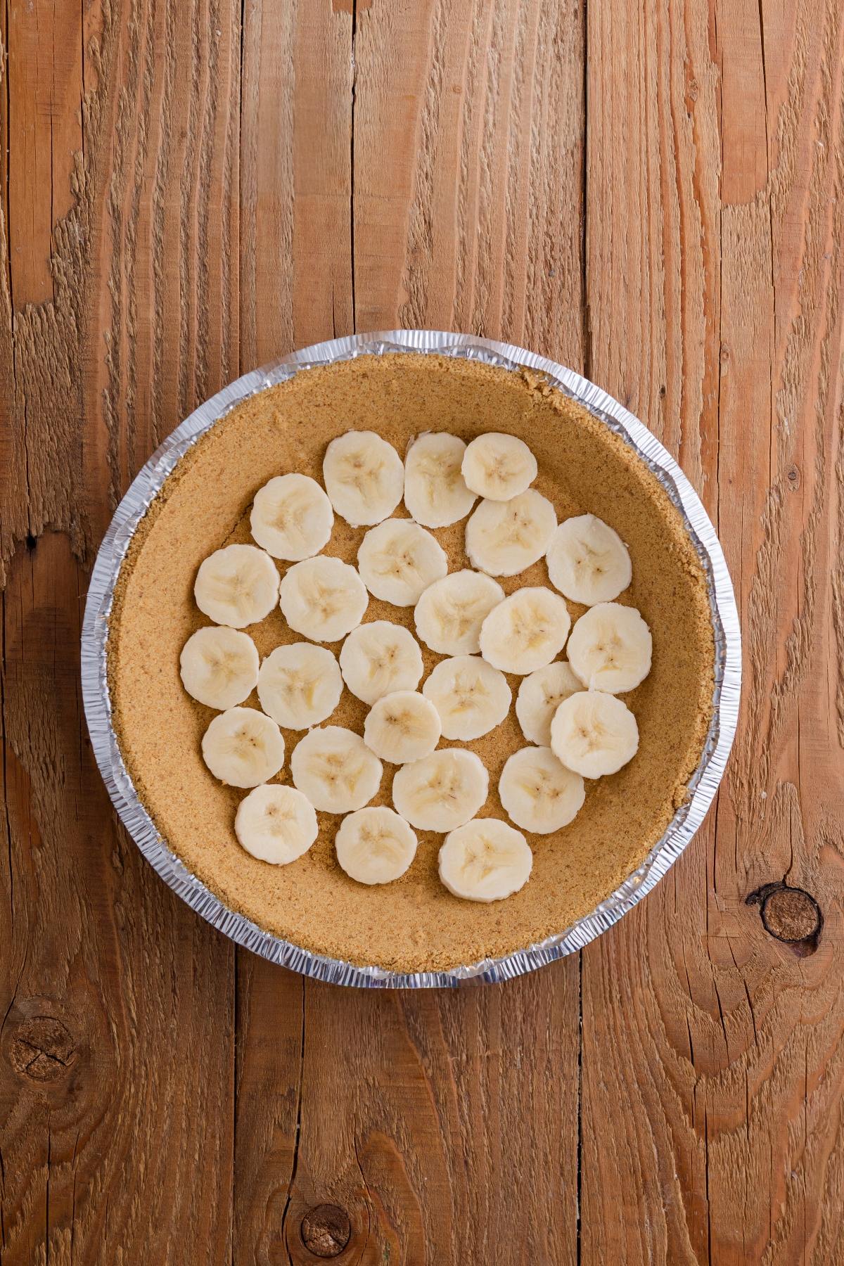 A pie crust filled with sliced bananas arranged in a single layer, on a wooden surface.