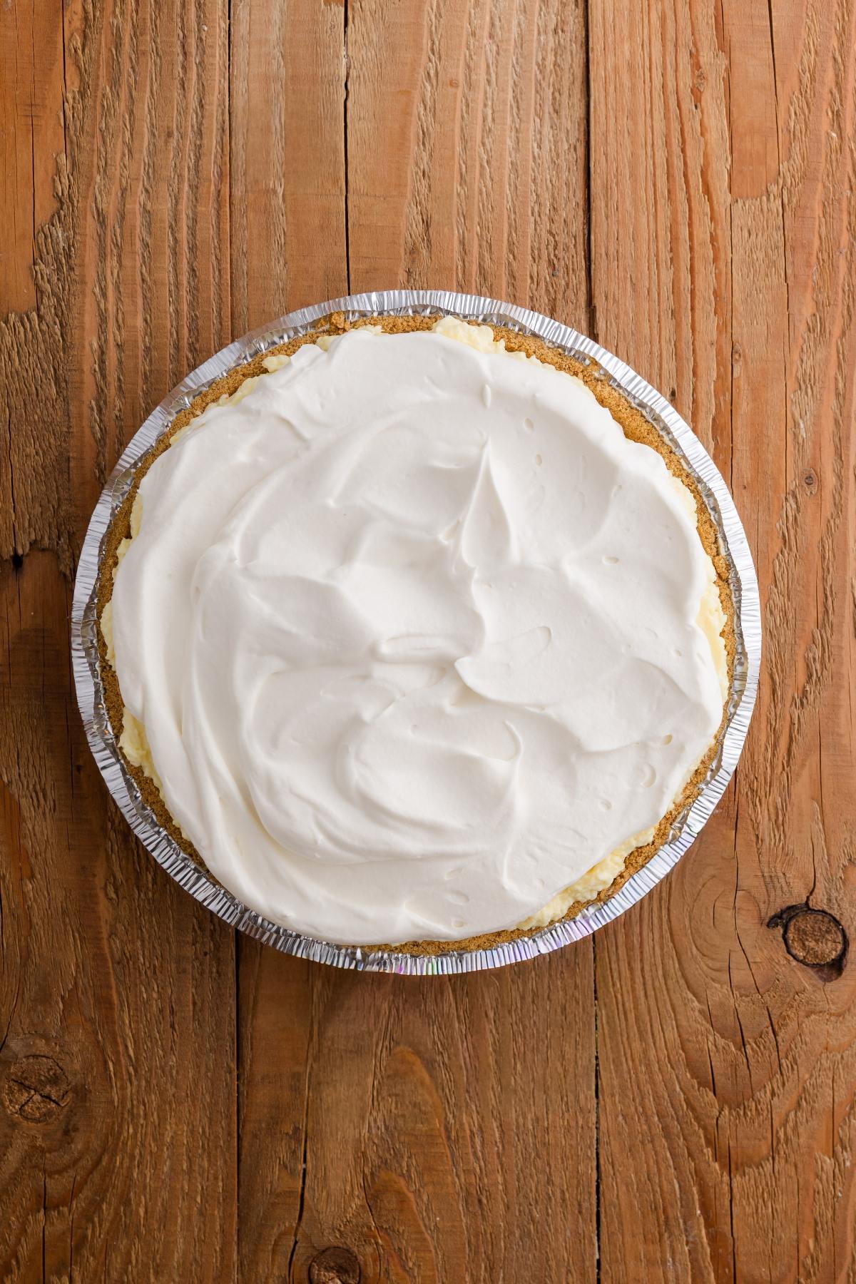 A cream pie with a whipped topping in a foil pan, placed on a wooden surface.