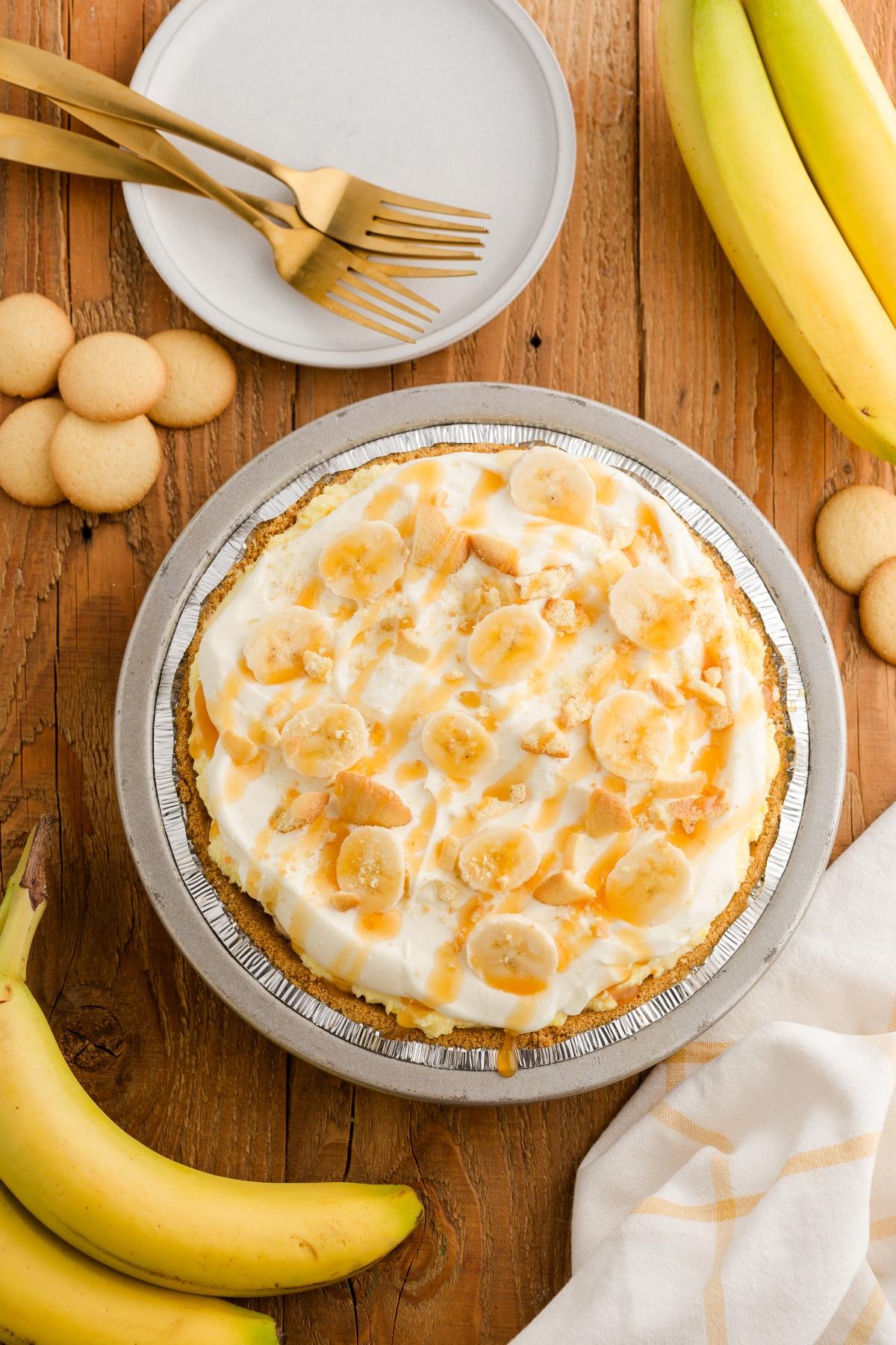A banana pudding pie topped with banana slices and wafers, surrounded by bananas and cookies on a wooden table.